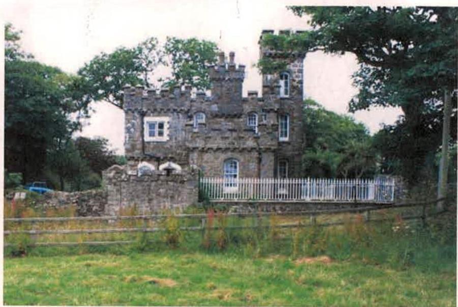 A photograph of a distinctive stone dwelling with castle-like crenellations, surrounded by mature trees and a stone wall with a white picket fence.