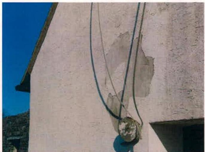 A close-up photograph of a rendered exterior wall showing a patch of exposed substrate and cables running down the surface. A stone fixture, possibly a pulley, is attached to the wall.