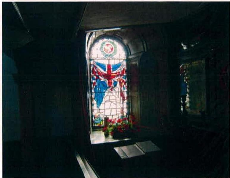 A photograph showing the interior of a historic building featuring a large arched stained glass window with a Union Jack design. Floral tributes are placed at the base of the window, illuminated by natural light.