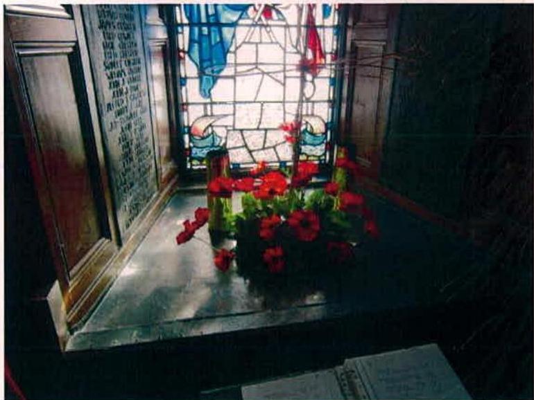 A photograph of an interior memorial featuring a stained glass window, a wooden board with inscriptions, and red flowers on a stone plinth.