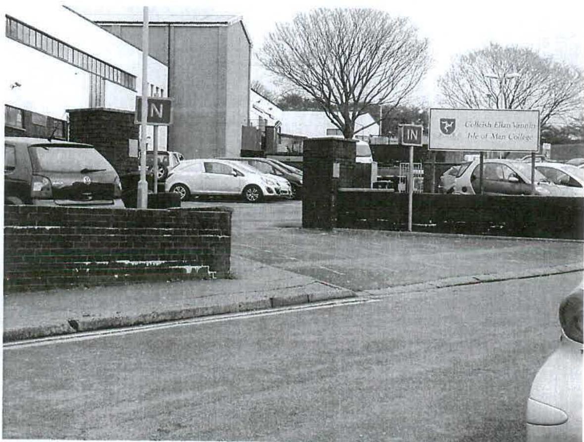 A black and white photograph showing the entrance to the Isle of Man College, featuring a brick boundary wall and parked cars.