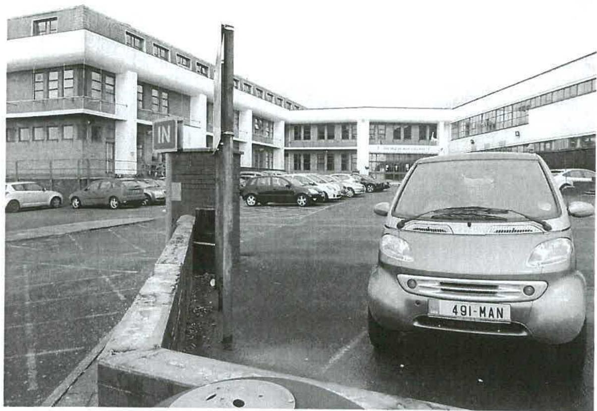 A black and white photograph showing a parking area entrance with a low brick boundary wall in the foreground and a large multi-story building in the background.