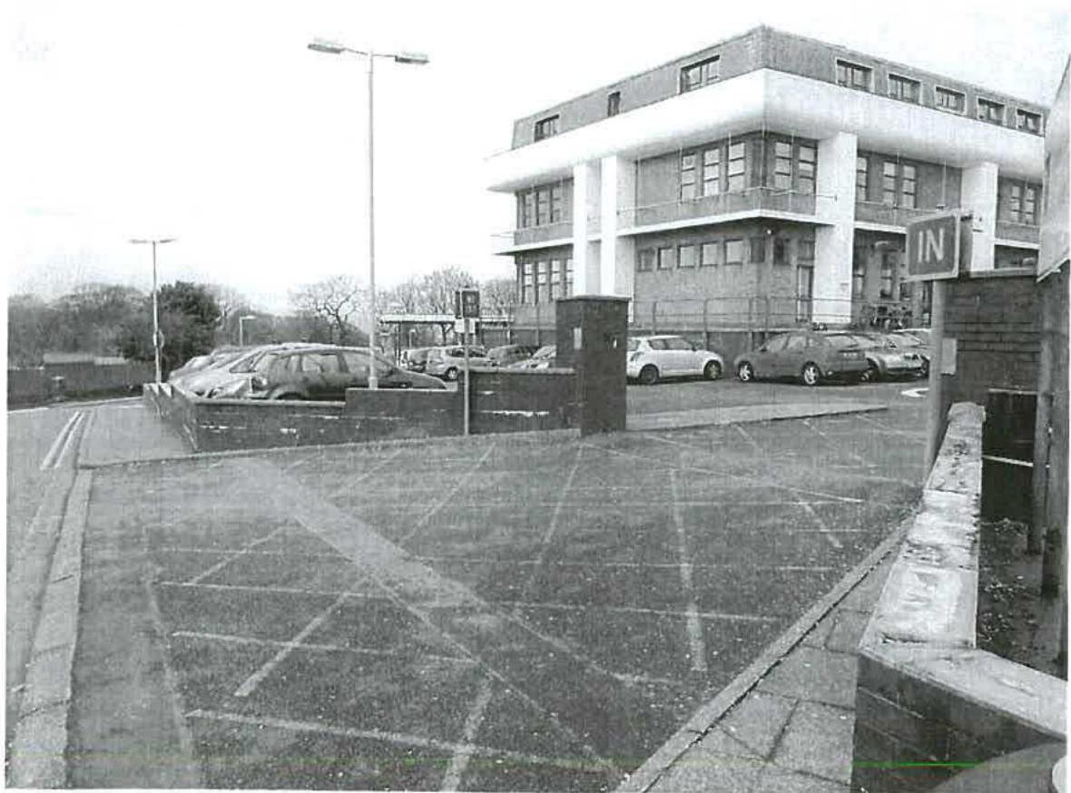 A black and white photograph showing a paved parking area with cars, a low boundary wall, and a multi-story commercial building in the background.