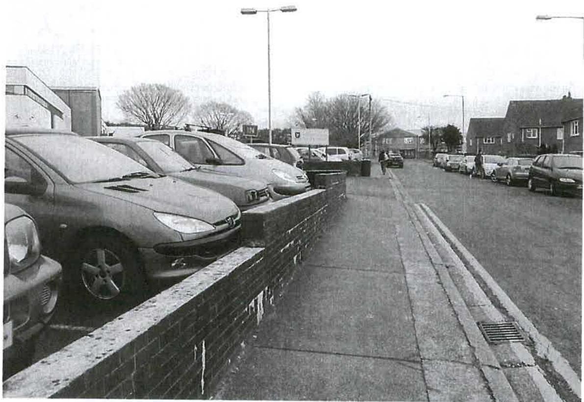 A black and white photograph showing a street scene featuring a low brick boundary wall in the foreground with parked cars behind it and a sidewalk running alongside.