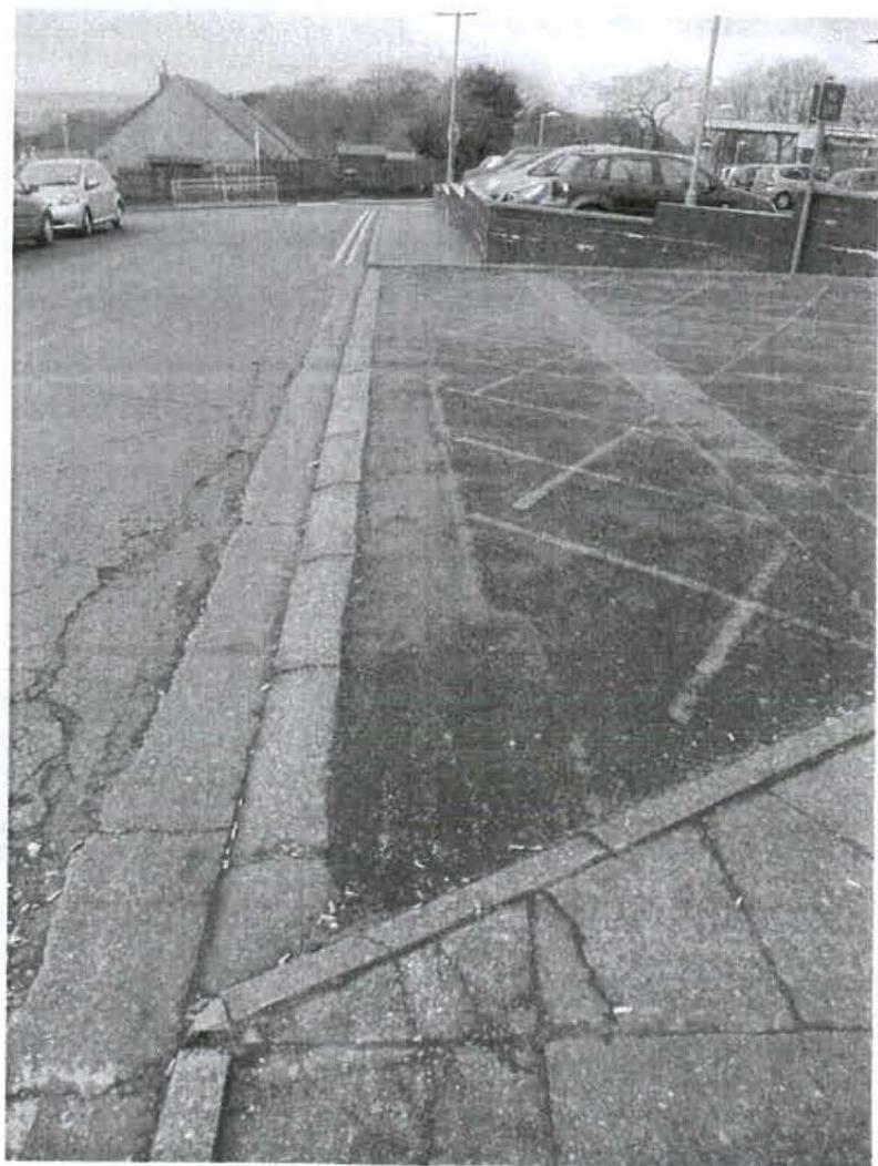 A black and white photograph showing the existing paved surface, curb, and parking markings at the site entrance or boundary area.