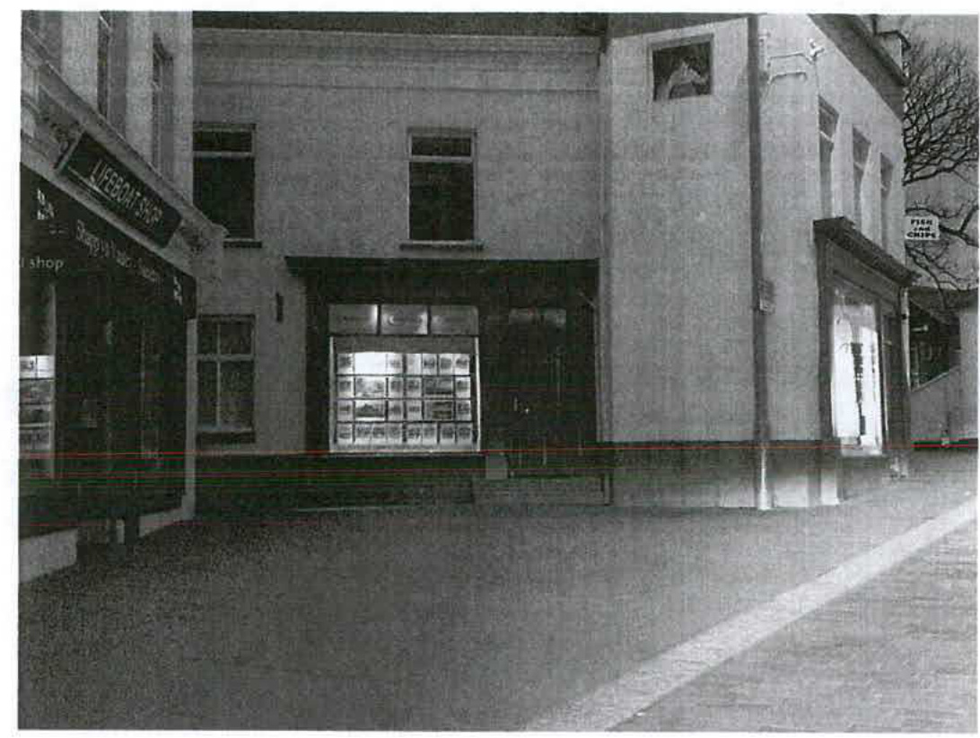 A grainy black and white photograph showing a street-level view of a row of commercial buildings, including a shop with a 'Livebonit Shop' sign and a 'Fish and Chips' shop.
