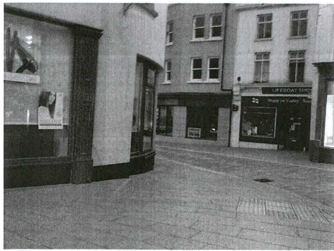 A black and white photograph showing a paved high street scene with shop fronts, likely serving as a context view for the proposed sculpture location.