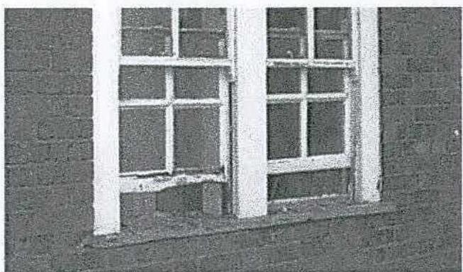 A grainy, black and white photograph showing a close-up of a white sash window on a dark wall. The window appears to have some damage or repair work at the bottom left section.