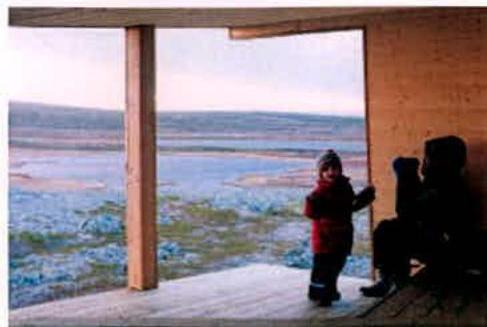 A photograph showing an interior view from a wooden structure looking out through a large opening onto a coastal landscape with a child standing nearby.