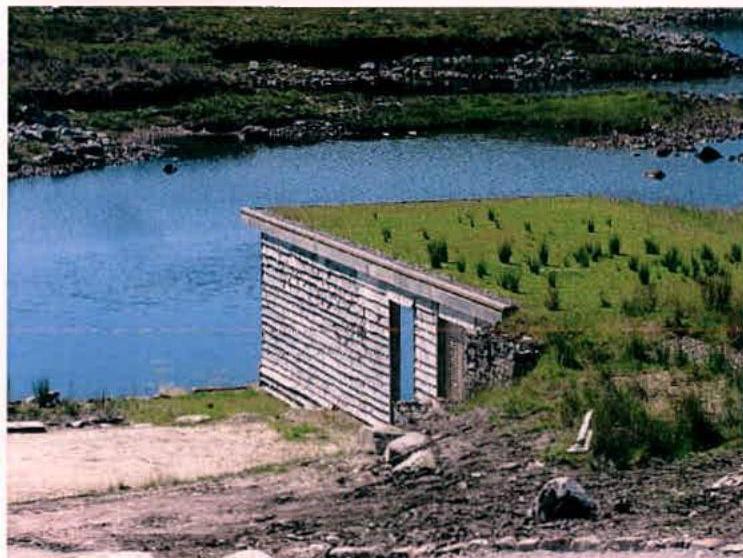 A photograph showing a small structure with a green roof and wooden cladding situated next to a body of water, likely representing the proposed new extension or outbuilding.