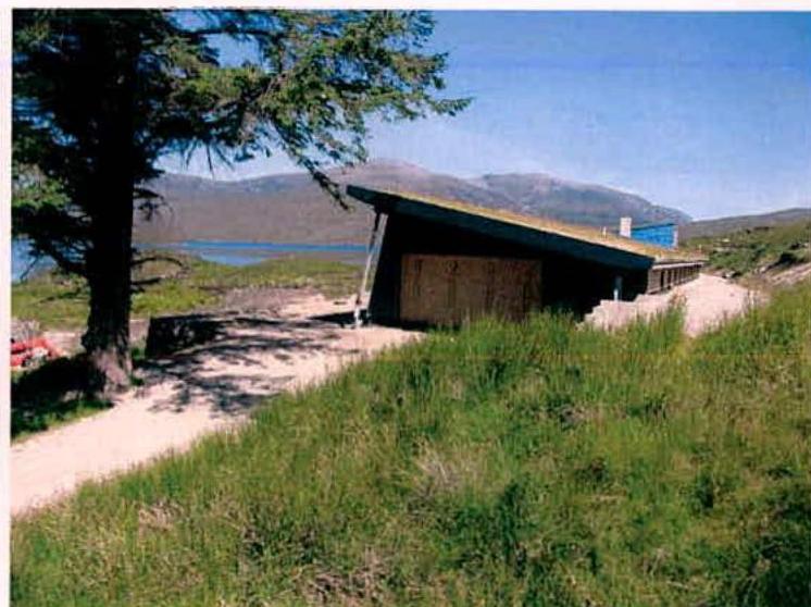 A photograph showing a wooden outbuilding or garage situated in a grassy, rural area with a large tree to the left and hills/water in the background.