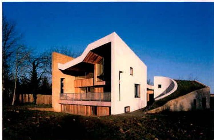 A photograph showing a modern, white-walled building with wooden cladding and a distinctive curved roofline, situated in a rural setting with a grassy mound to the side.