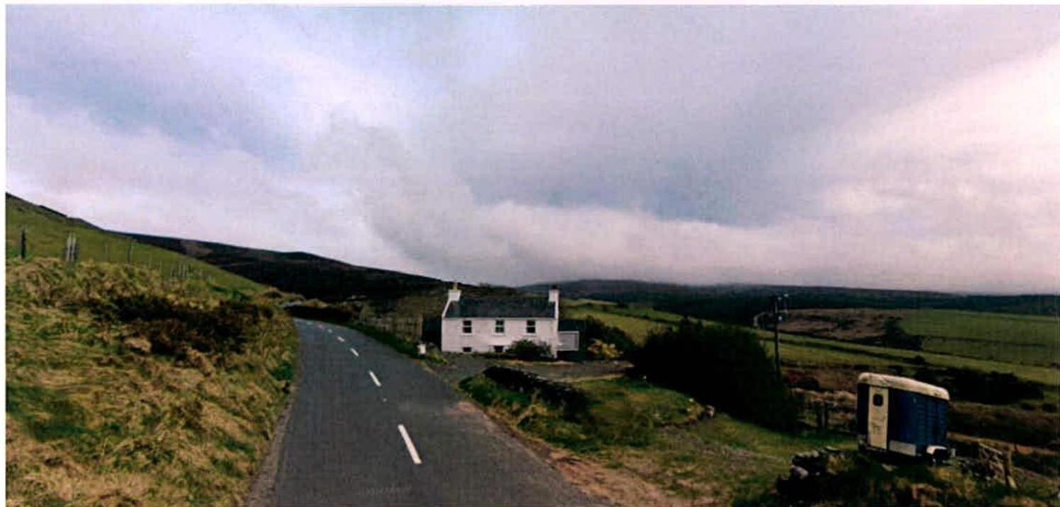 A white detached cottage situated on a grassy hillside next to a winding road, with a portable toilet visible in the foreground suggesting construction activity.