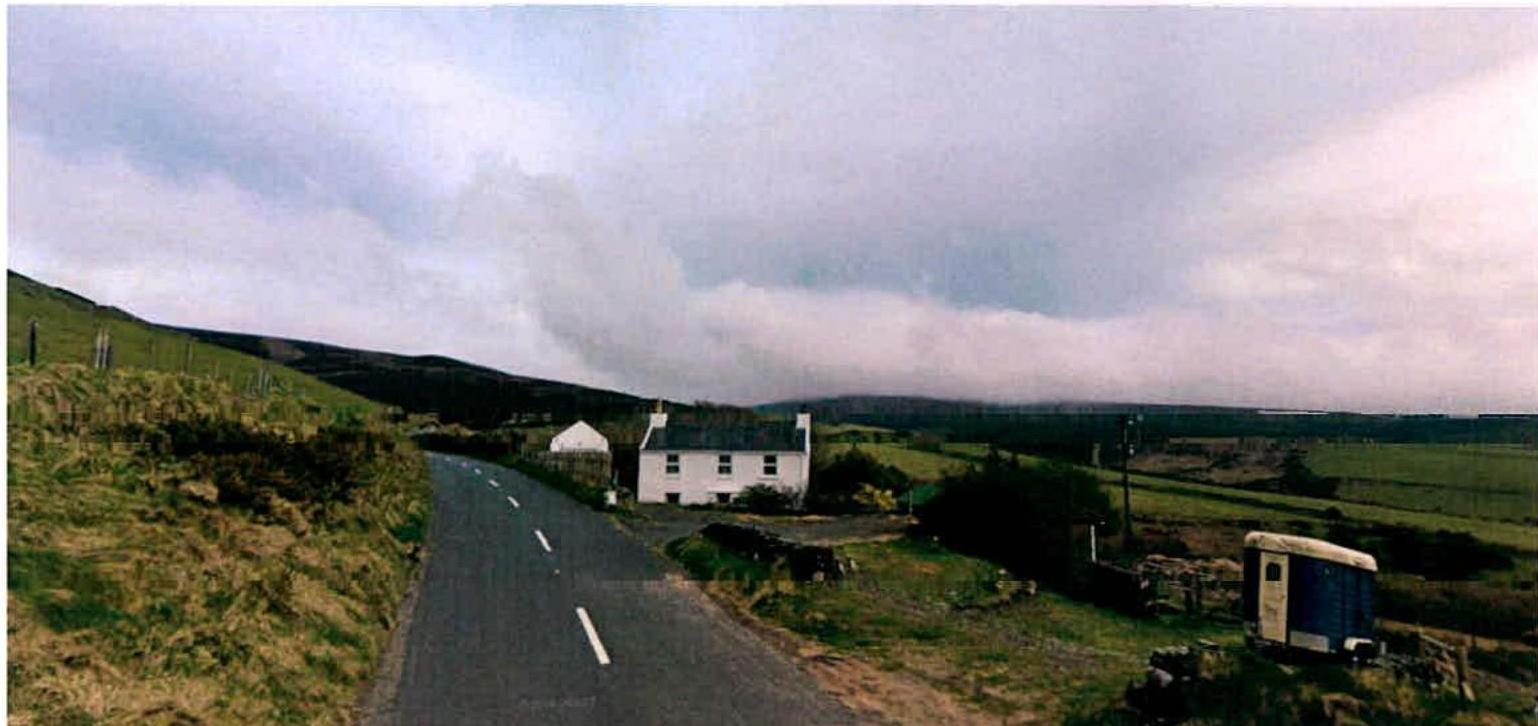 A photograph showing a white detached cottage situated alongside a rural road with green hills in the background. The image captures the existing site conditions including a small outbuilding and a caravan.