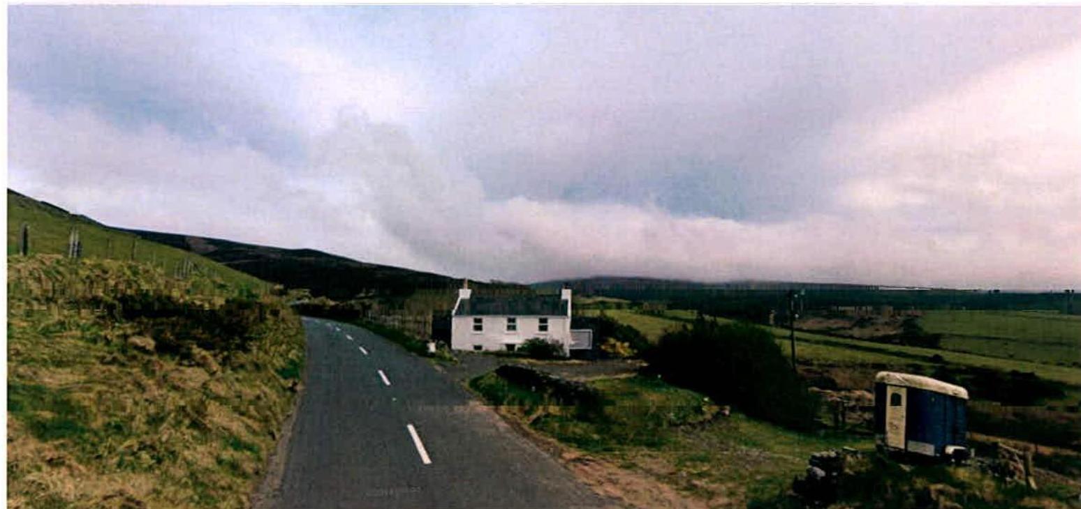 A wide-angle photograph showing a white two-story cottage situated beside a rural road with green hills in the background.
