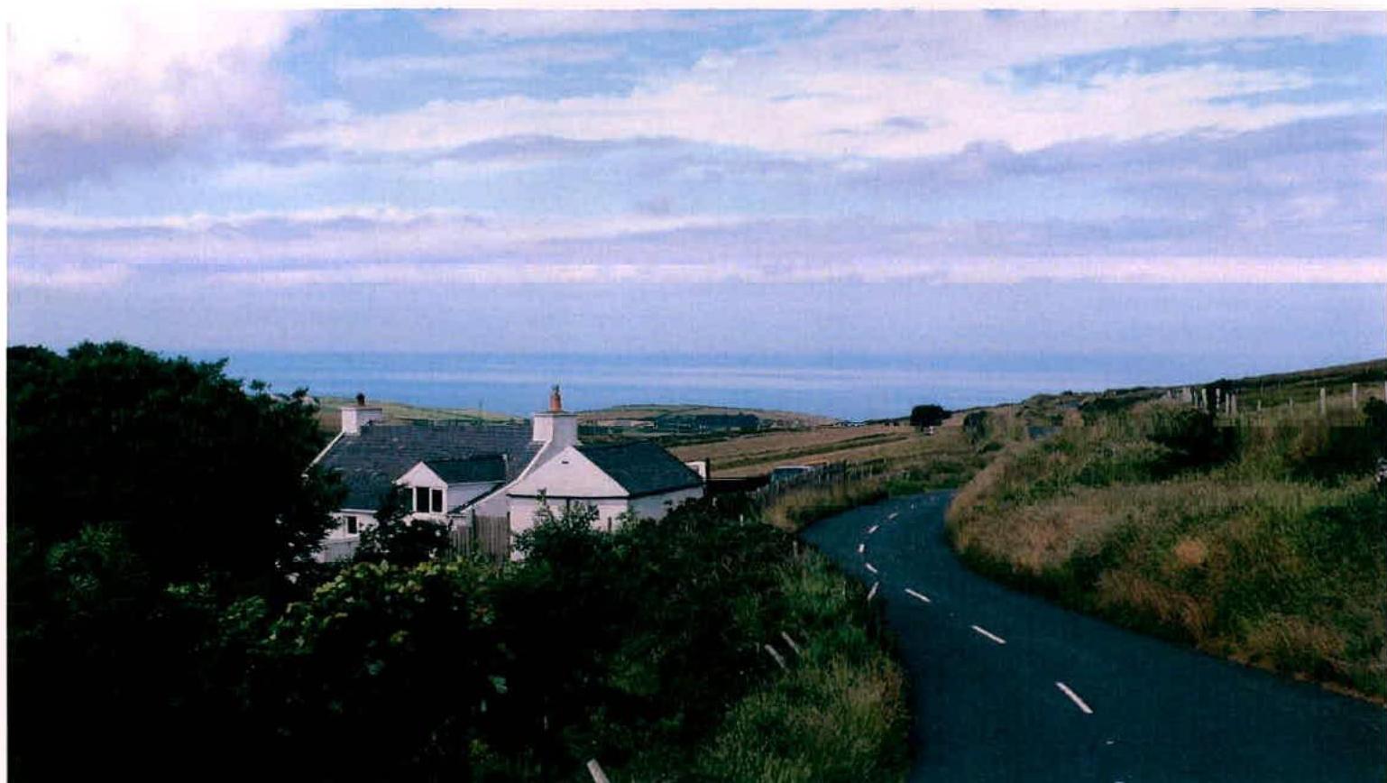 A photograph showing a white cottage situated on a grassy hillside with a winding road in the foreground and the sea visible in the distance.