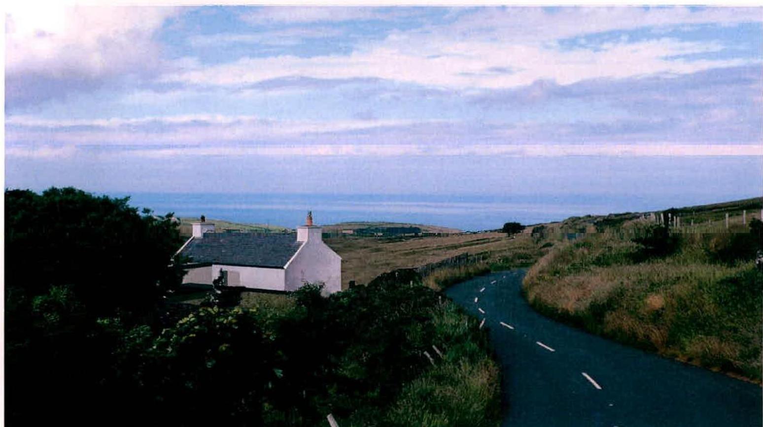 A landscape photograph showing a white cottage situated on a grassy hillside next to a winding road, with the sea visible in the background.
