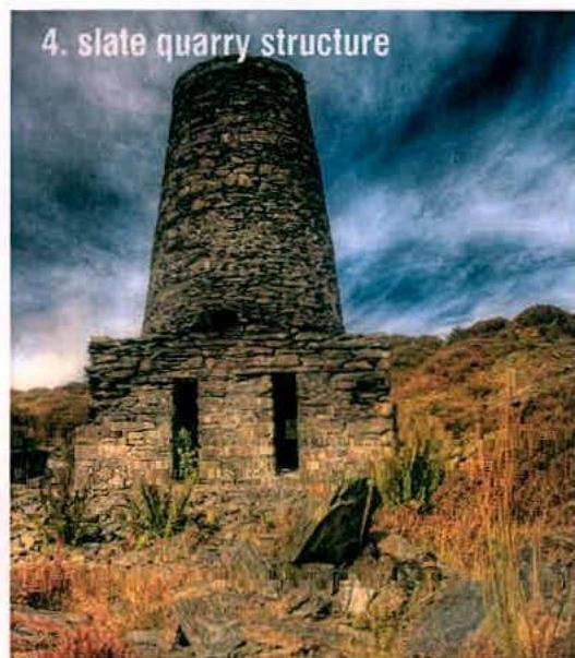 A photograph of a stone slate quarry structure, appearing as a tall tower with a rectangular base, set against a cloudy sky and rural hillside.