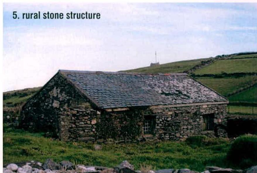 A photograph showing a single-story rural stone building with a slate roof, set against a grassy hillside with stone walls.
