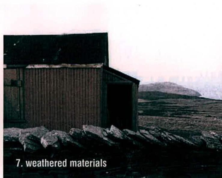 A photograph showing a weathered corrugated outbuilding or garage with a stone wall in the foreground and a coastal landscape in the background.