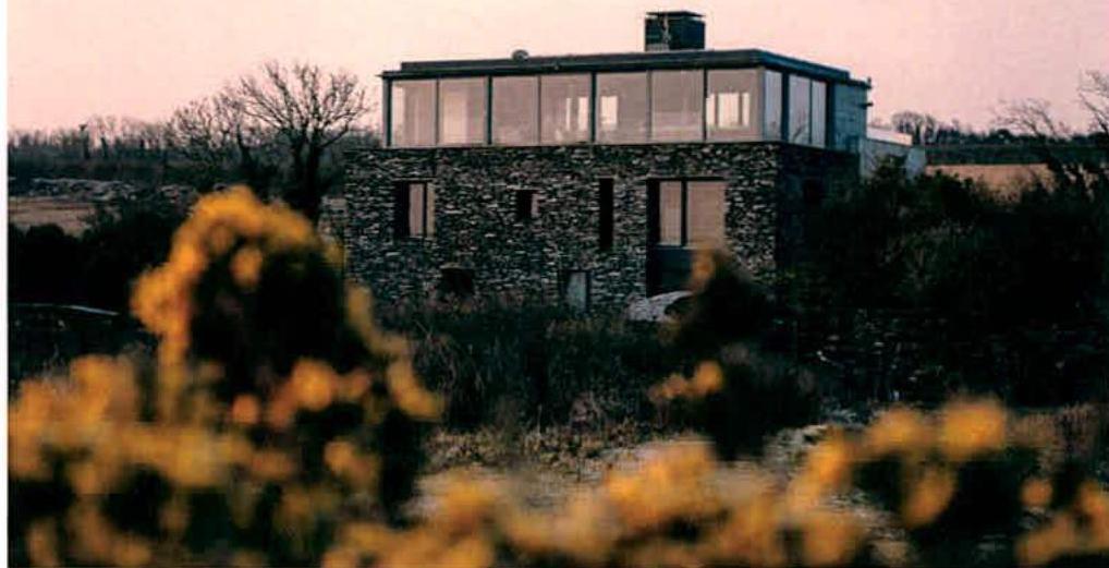 A photograph showing a two-story stone building with a modern glass upper extension, situated in a rural landscape with gorse in the foreground.