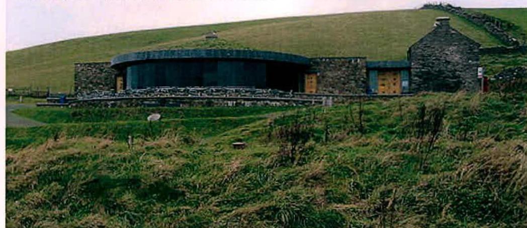 A photograph showing a rural building complex featuring a traditional stone structure on the right and a large modern extension with a curved roofline on the left, set against a grassy hillside.