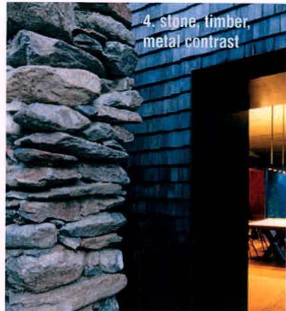 A close-up architectural photograph showing a rough stone wall adjacent to dark cladding and a doorway leading to a lit interior.