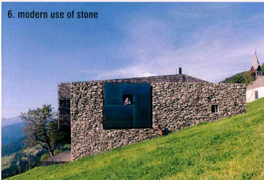 A photograph of a modern stone building situated on a grassy slope with a church spire visible in the background.