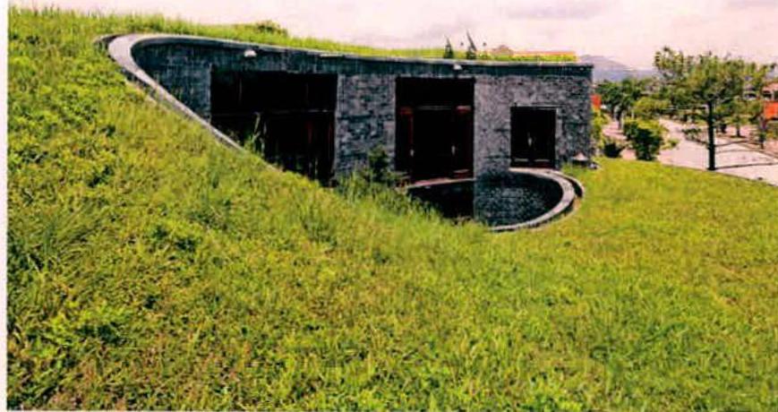 A photograph showing a modern building with a curved stone facade integrated into a grassy hillside, featuring dark openings and a green roof.