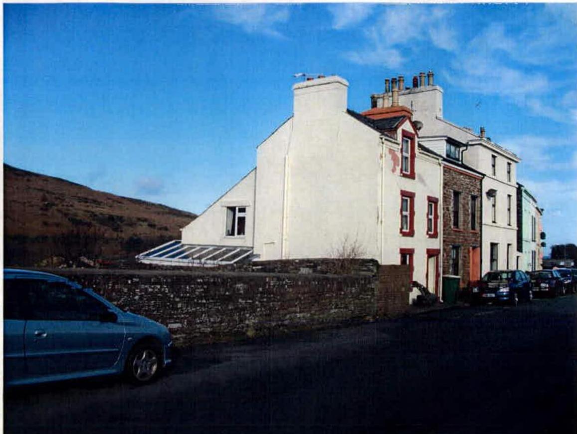 A street-level photograph showing a white two-storey building adjacent to a row of terraced houses, with a hill in the background and cars parked on the street.
