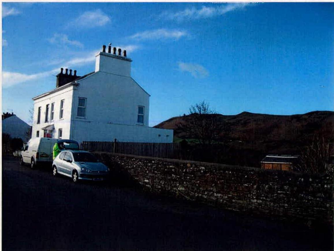 A photograph showing a white two-story building with a stone wall in the foreground and a hill in the background, with vehicles parked on the road.