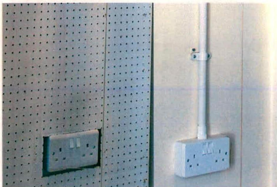 A close-up photograph of an interior wall featuring a perforated pegboard panel with a socket and an adjacent smooth wall with a vertical electrical conduit and socket.