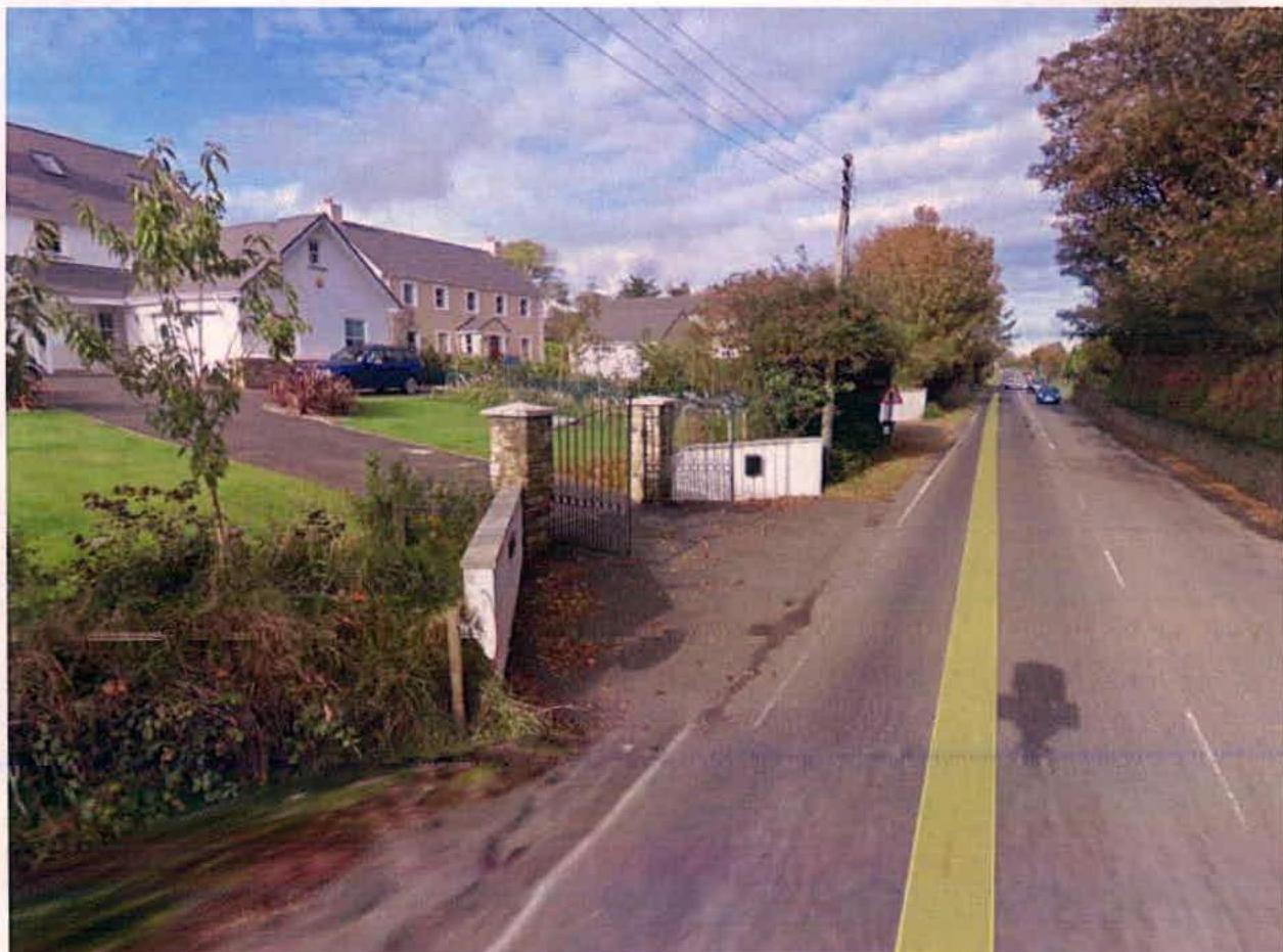 A street-level photograph showing a white detached house with a gated entrance and driveway adjacent to a road with a yellow center line.