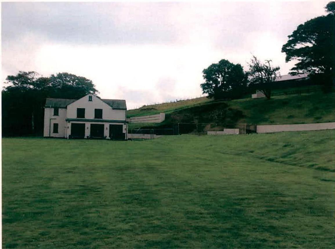 A photograph showing a white detached building with three garage doors situated next to a large grassy field and a sloping hill.