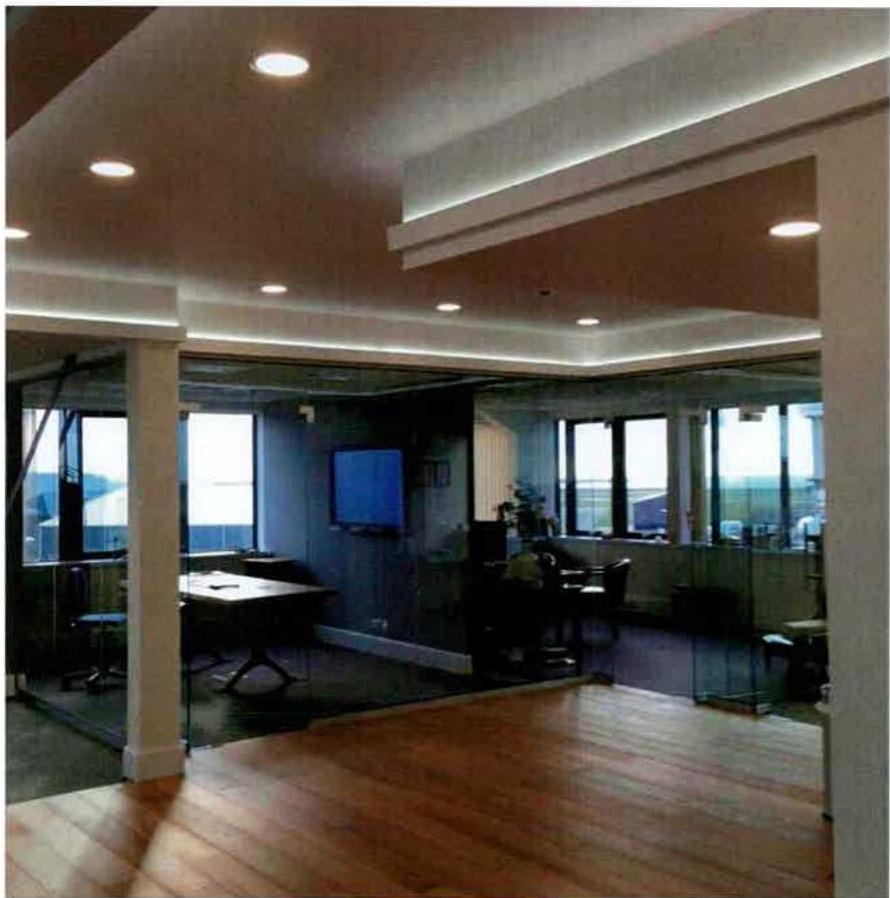 Interior photograph of a modern office space featuring wooden flooring, glass partitions, and recessed ceiling lighting.