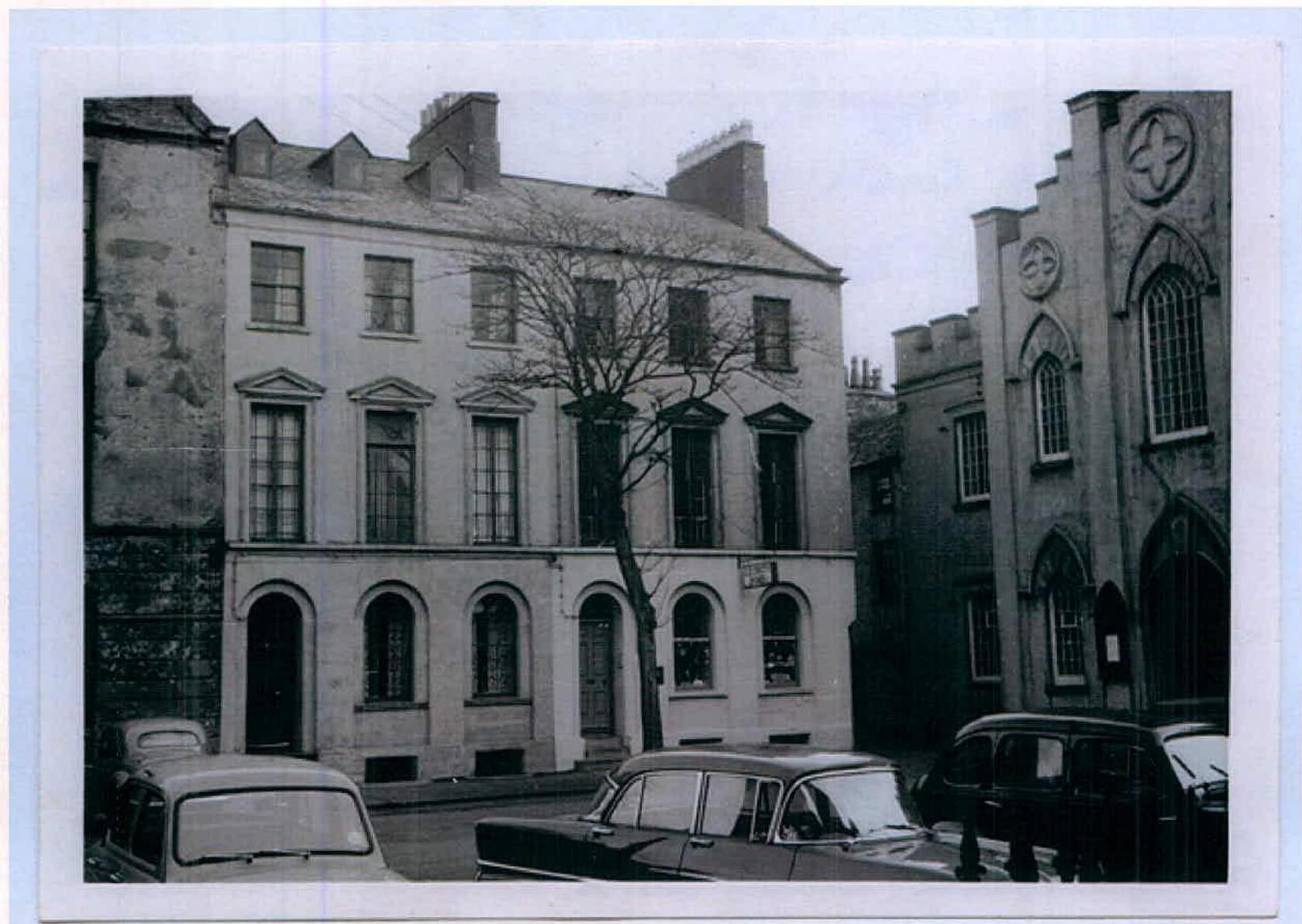 A black and white photograph showing a historic street scene featuring a large multi-story building with arched ground-floor windows and sash windows above, alongside a Gothic-style building.