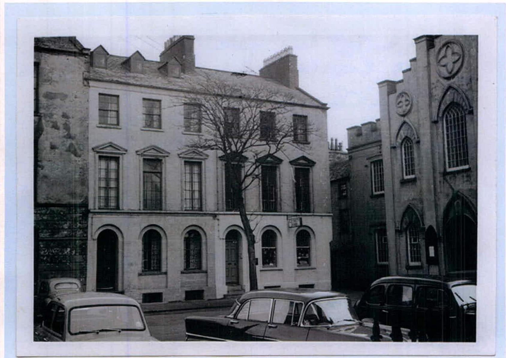 A black and white photograph showing a three-story building with classical architecture, likely a former office, situated next to a Gothic-style building.