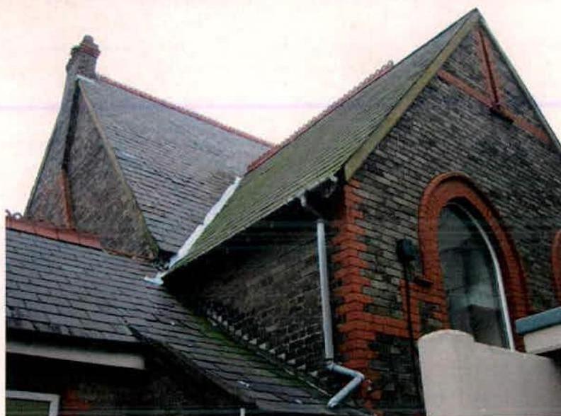 A photograph showing the upper facade and roofline of a brick building with slate roofing and red brick detailing around an arched window.