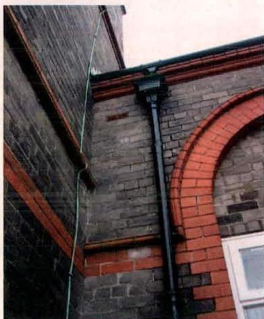 A close-up photograph of a brick building exterior featuring a red brick archway, grey brickwork, and a vertical black drainpipe.