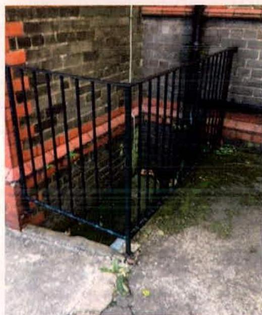 A photograph showing a black metal railing installed around a sunken brick area, likely a basement lightwell or courtyard, adjacent to a brick wall.