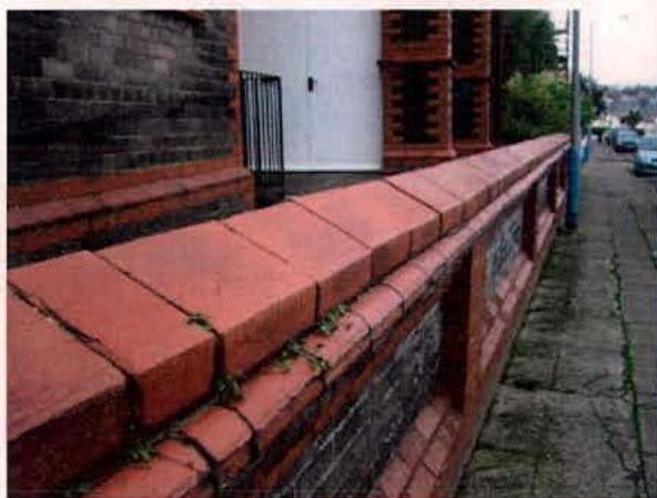 A low red brick boundary wall with coping stones runs along a paved sidewalk, with a black metal gate and white building facade visible in the background.