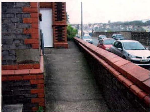 A photograph showing a brick boundary wall and paved walkway next to a street with parked cars.