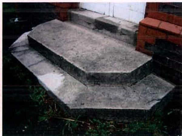 A close-up photograph showing weathered concrete steps or a ramp leading up to a brick building entrance.