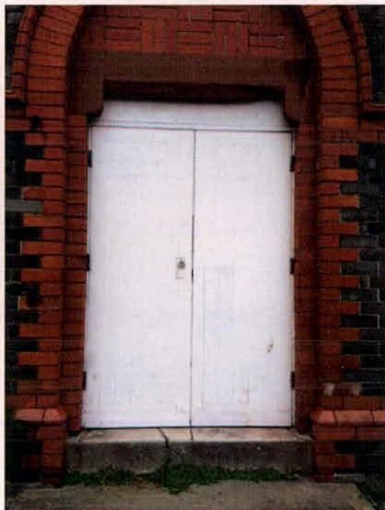 A close-up photograph showing a large white double door set within a decorative red brick archway with polychrome detailing. The image highlights the architectural detail of the entrance, likely part of a listed build...