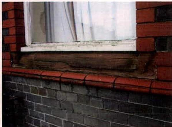 A close-up photograph showing a white window frame, a weathered wooden lintel, and red and grey brickwork.