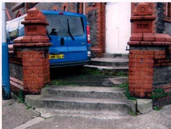 A photograph showing the entrance to a property featuring red brick gate pillars, stone steps leading to a white door, and a blue van parked behind the boundary.