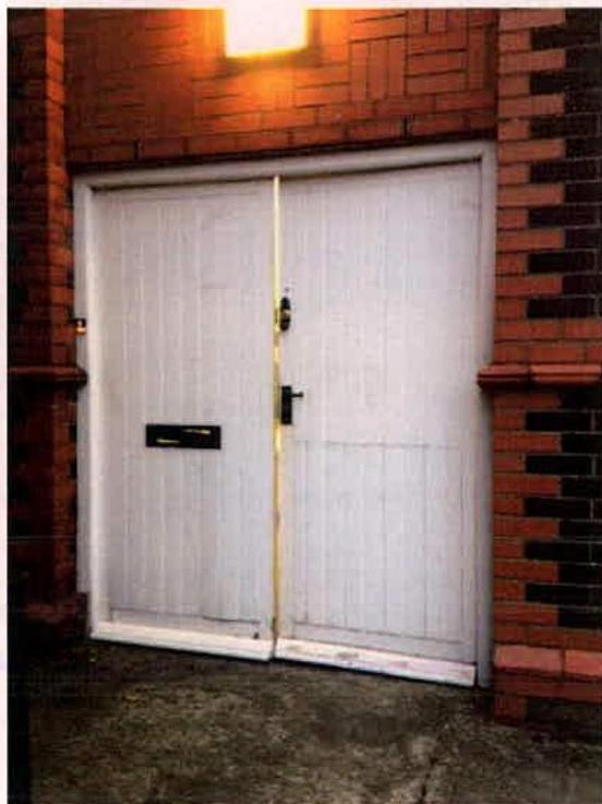 A close-up photograph of a white double door set within a red brick wall, featuring a mail slot and an overhead light fixture.