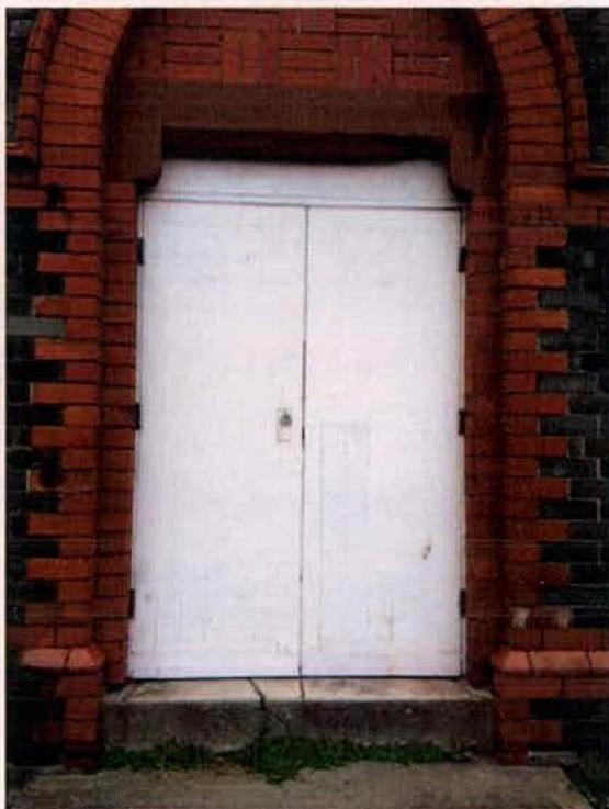 A close-up photograph of a large white double door set within a decorative red brick archway with black brick detailing.
