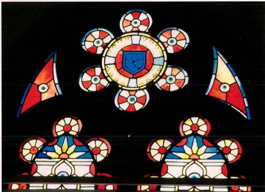 A close-up photograph of a stained glass window featuring a central blue shield with a triskelion surrounded by red and white floral patterns.