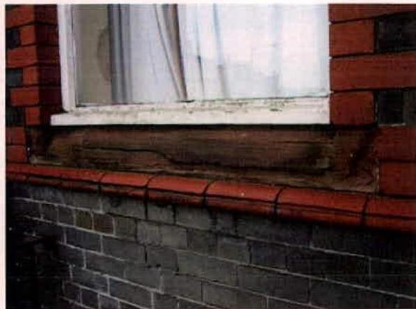 A close-up photograph of a window reveal showing a weathered wooden lintel, red brickwork, and a dark grey brick wall below.